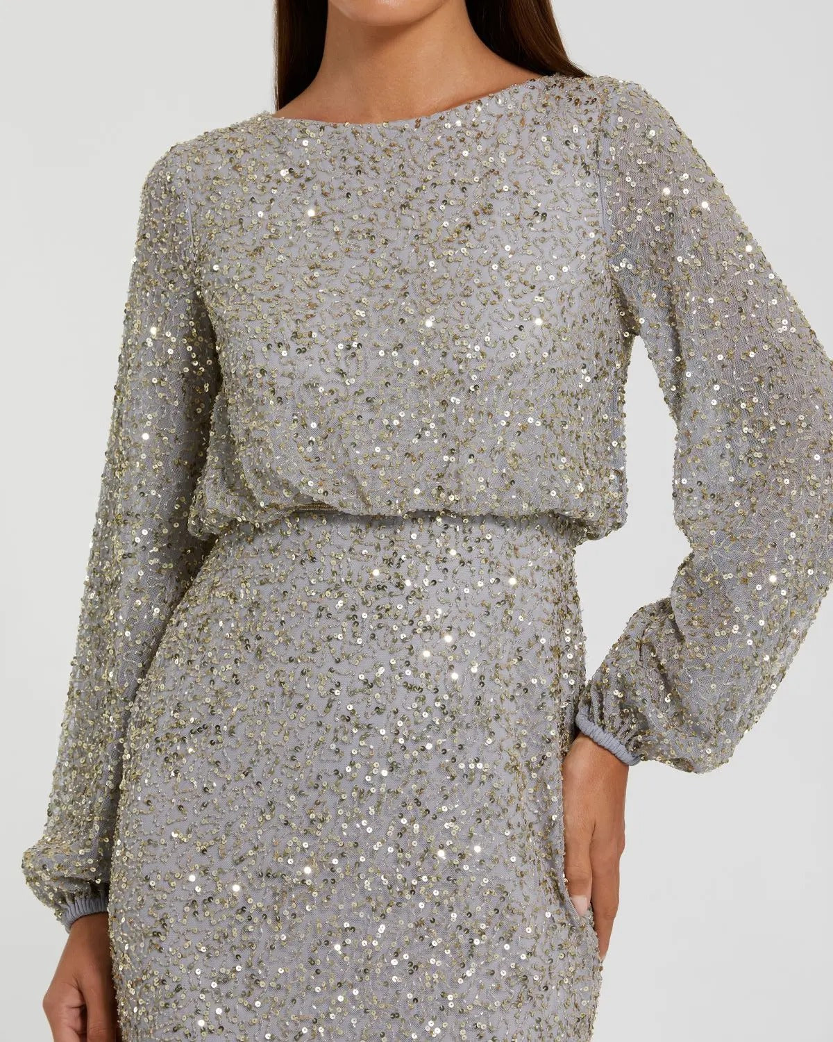 A person wearing the Mac Duggal 94203 long formal dress—a modest, long-sleeved gown in light gray adorned with shimmering silver and gold sequins—stands with one hand on their hip, embodying elegance and glamour.